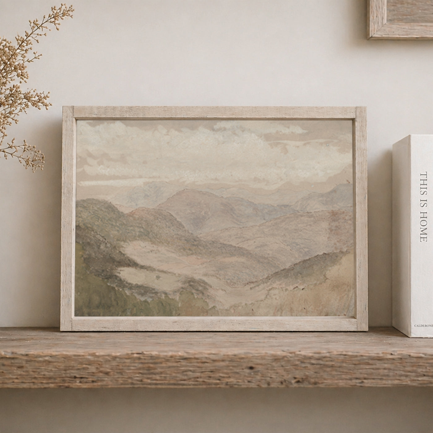 Framed mountain landscape painting on a wooden shelf with a book next to it.