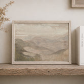 Framed mountain landscape painting on a wooden shelf with a book next to it.