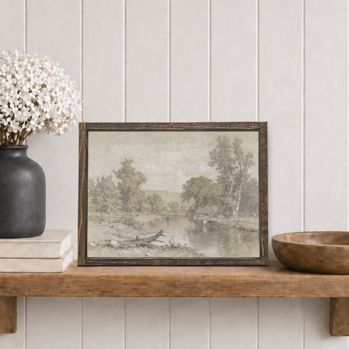 Framed landscape print  with a river and trees on a wooden shelf with decorative items against a white panelled wall.