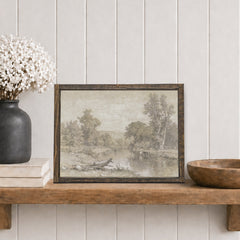 Framed landscape print  with a river and trees on a wooden shelf with decorative items against a white panelled wall.