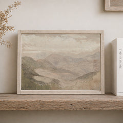 Framed mountain landscape painting on a wooden shelf with a book next to it.