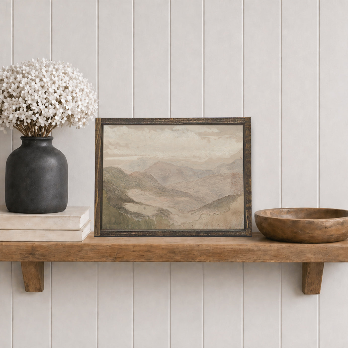 Framed soft mountain landscape print on a wooden shelf with a vase and bowl against a white paneled wall.