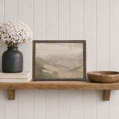 Framed soft mountain landscape print on a wooden shelf with a vase and bowl against a white paneled wall.