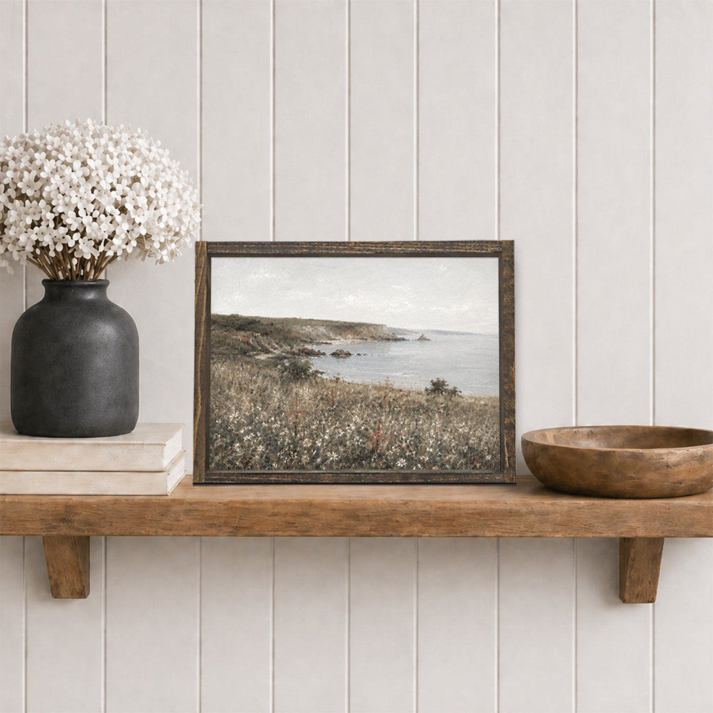 Framed landscape print on a wooden shelf with decorative items against a white paneled wall.