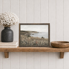 Framed landscape print on a wooden shelf with decorative items against a white paneled wall.