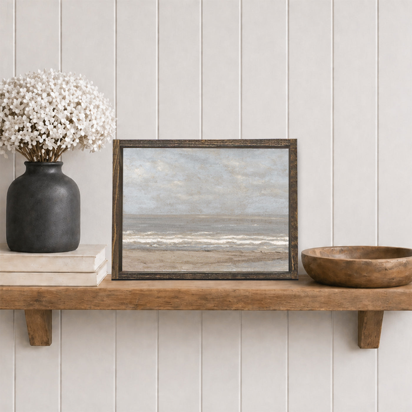 vintage ocean painting with espresso brown wood frame on shelf with a black vase with white flowers